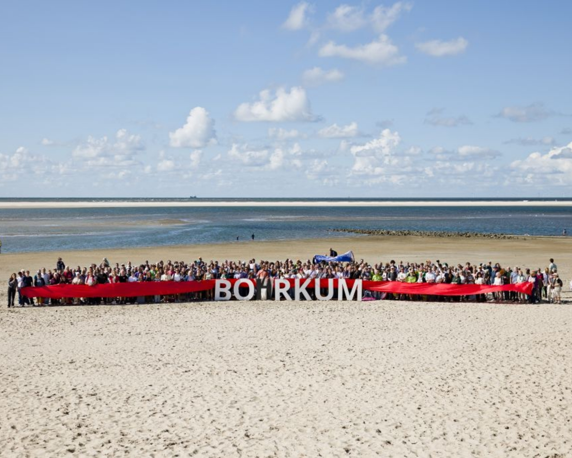 Eine Große Menge Menschen steht mit einem sehr großen Banner am Strand. Auf dem Banner steht Bohrkum in Großbuchstaben (Der Name der Insel Borkum, mit einem zusätzlichen H, ein Wortspiel mit dem Wort bohren), wobei das H gleichzeitig eine Zeichnung einer Bohrinsel ist.